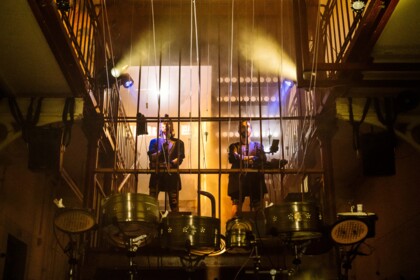 Eine Performance in Zellentrakt der JVA Rennelberg.  Zwei Performer:innen stehen auf einer Treppe hinter Metallstangen. Gelbes Bühnenlicht und Nebel erzeugen eine dichte Atmosphäre. Im Vordergrund auf dem Boden ist ein Schlagzeug aufgebaut. 